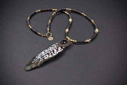 A bohemian-style beaded necklace featuring a large feather pendant, black and gold beads, and a central dark stone, displayed on a dark gray background.