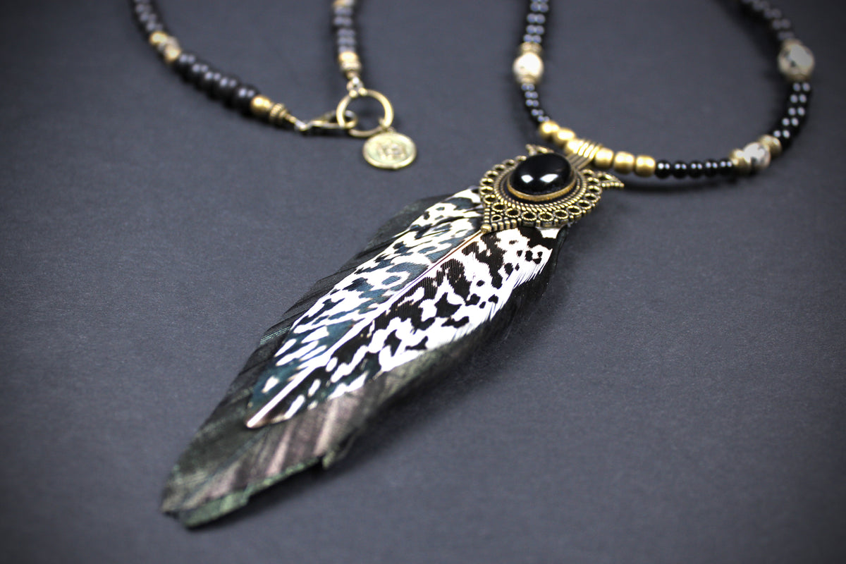 A bohemian-style beaded necklace featuring a large, realistic black and white patterned feather pendant with a black cabochon and gold-toned filigree setting, displayed on a dark gray background.