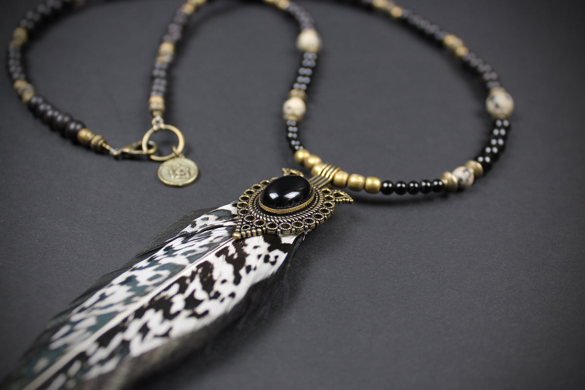 A bohemian-style beaded necklace featuring a large black and white patterned feather pendant, set in an ornate bronze-toned metal frame with a black cabochon. The necklace is composed of black beads, gold-toned beads, and white stone beads, with a small gold charm engraved with VB