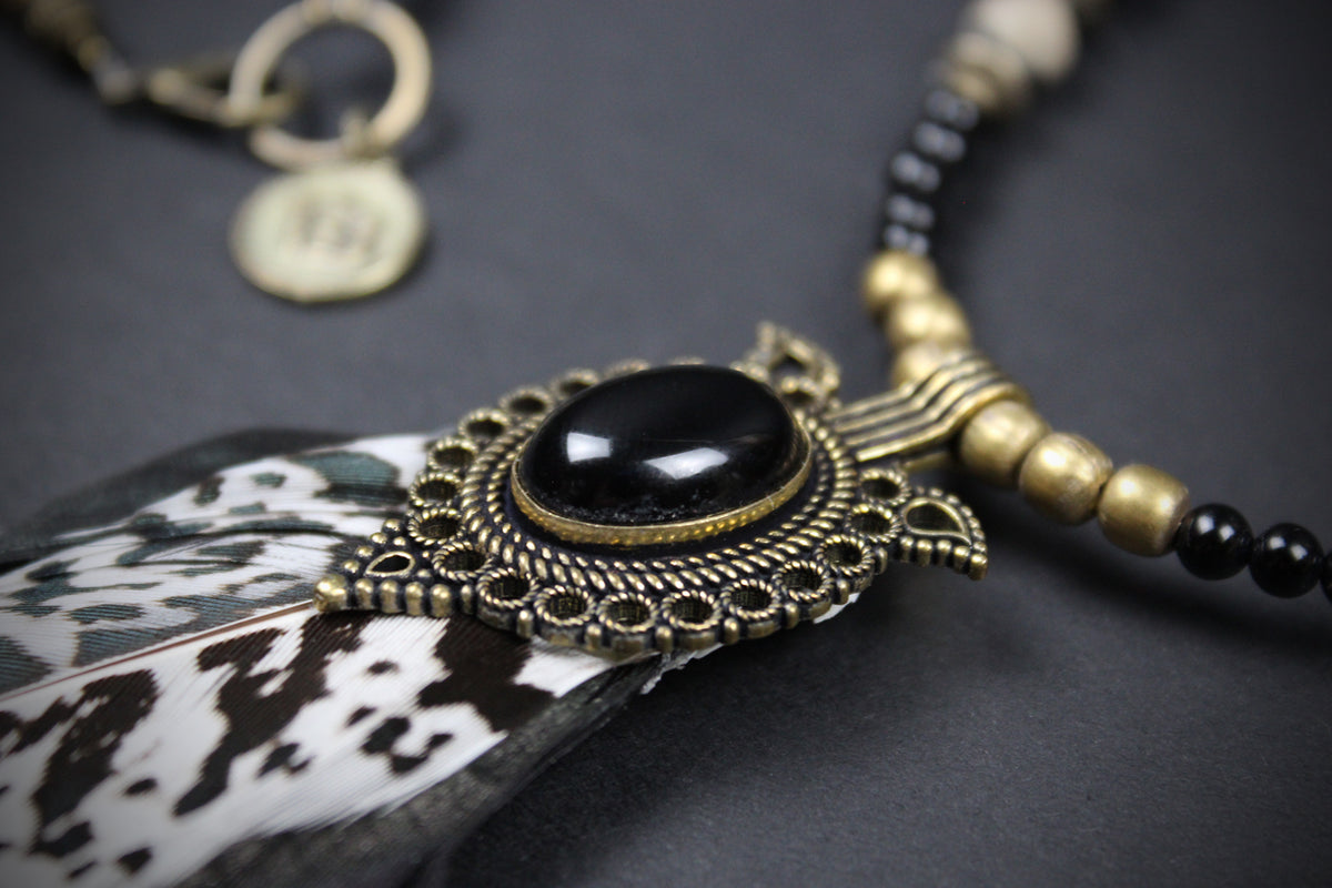 A close-up of a bohemian-style necklace featuring a black cabochon stone in an ornate antique gold-toned filigree frame, resting on a black and white cowhide feather. The necklace also includes black and gold beads and a small round tag.