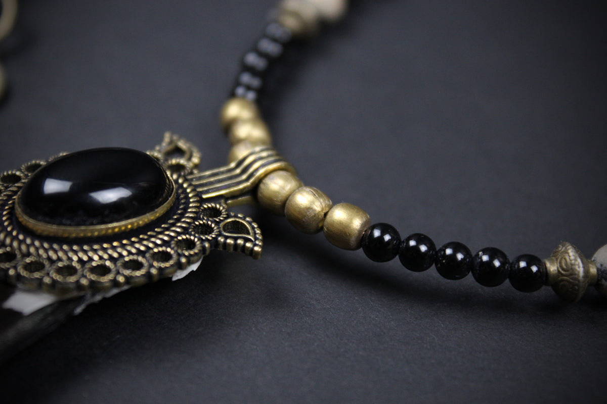 A close-up of a beaded necklace featuring a large oval black stone in an ornate antique bronze filigree setting, accented by small black beads and matte bronze cylindrical spacers on a dark background.