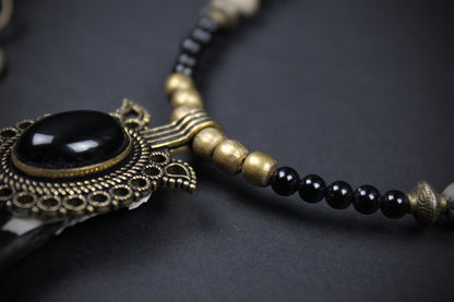 A close-up of a beaded necklace featuring a large oval black stone in an ornate antique bronze filigree setting, accented by small black beads and matte bronze cylindrical spacers on a dark background.