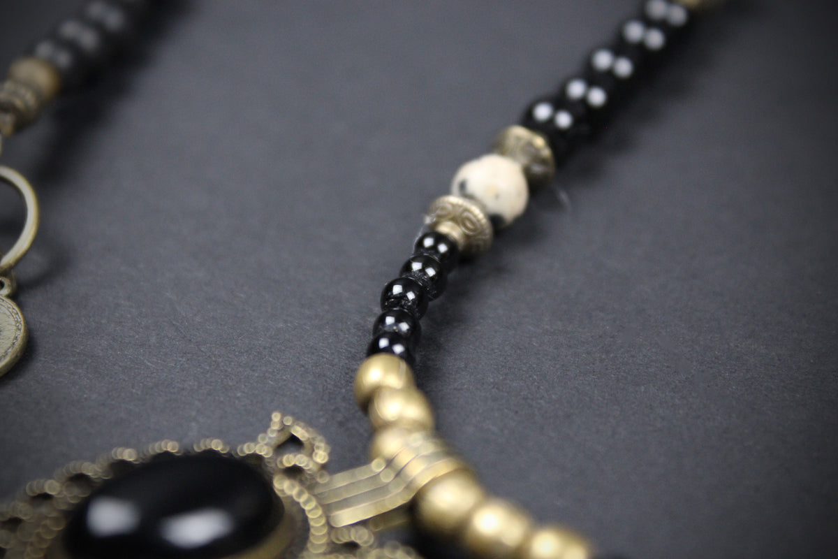 Close-up of a beaded necklace with black round beads, a white bead, antique gold-toned metal spacers, and a large black oval stone pendant on an ornate gold-toned frame, set against a dark gray background.
