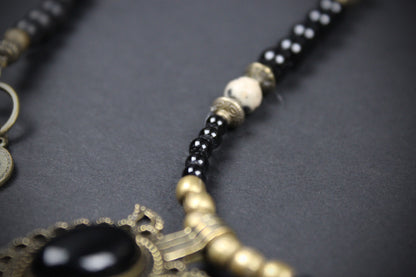 Close-up of a beaded necklace with black round beads, a white bead, antique gold-toned metal spacers, and a large black oval stone pendant on an ornate gold-toned frame, set against a dark gray background.