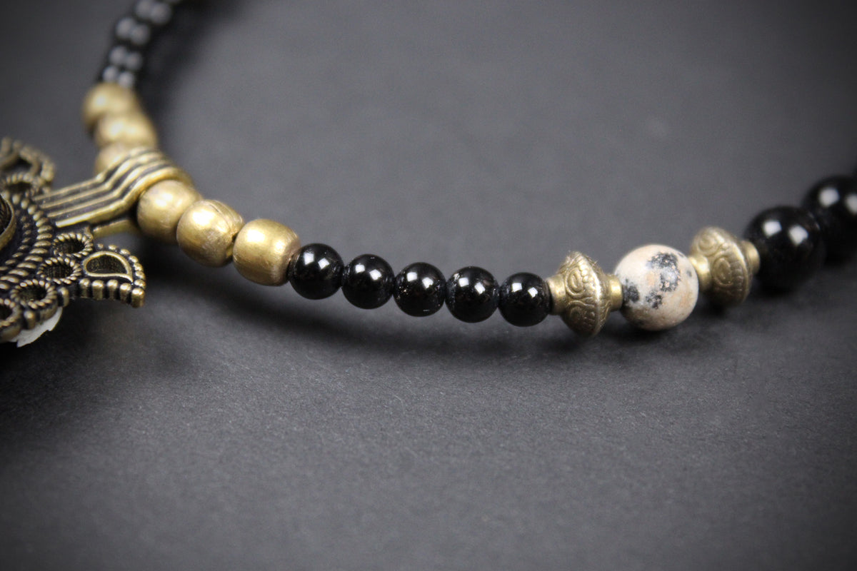 A close-up of a beaded necklace featuring glossy black round beads, antique gold metallic spacer beads, a white marble accent bead with black veining, and a detailed antique gold pendant on a dark gray background.