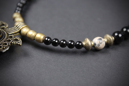 A close-up of a beaded necklace featuring glossy black round beads, antique gold metallic spacer beads, a white marble accent bead with black veining, and a detailed antique gold pendant on a dark gray background.