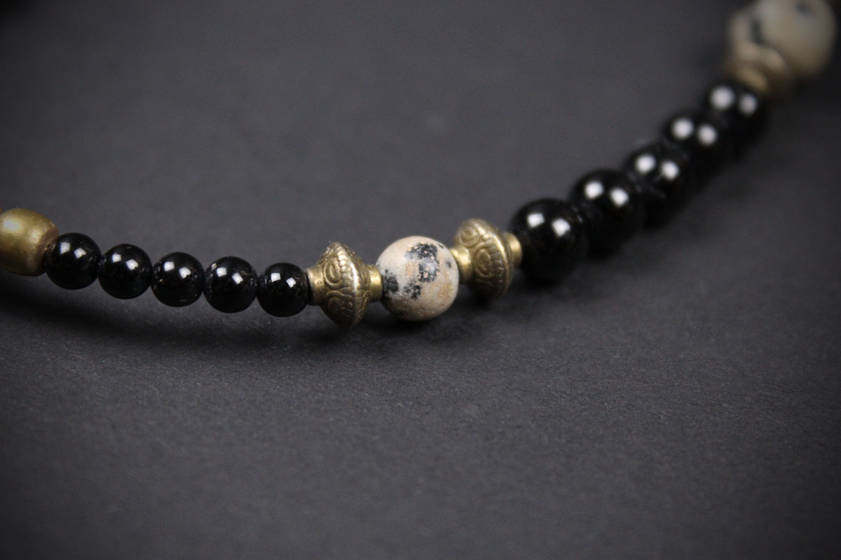 A close-up of a beaded necklace featuring glossy black round beads, antique bronze metallic spacers, and a central round white bead with black marbled patterns, set against a dark gray background.