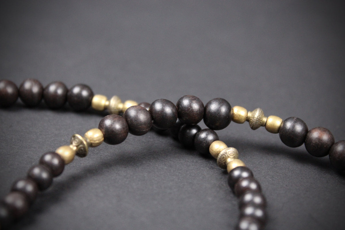 Dark brown wooden bead necklace with small metallic gold spacer beads, shown in a close-up shot against a dark gray background.