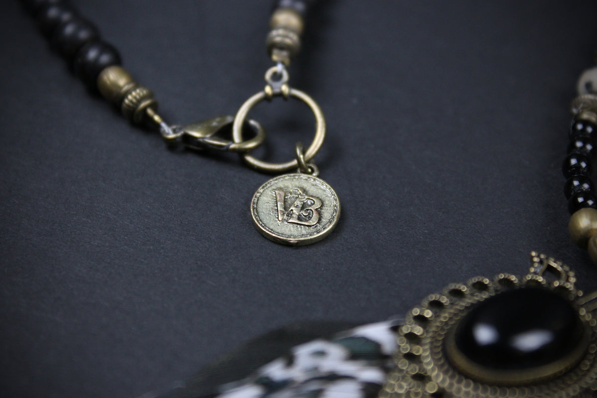 Close-up of a beaded necklace with black beads, antique bronze-toned metal spacers, a ring clasp, and a round coin-style pendant on a dark textured background.