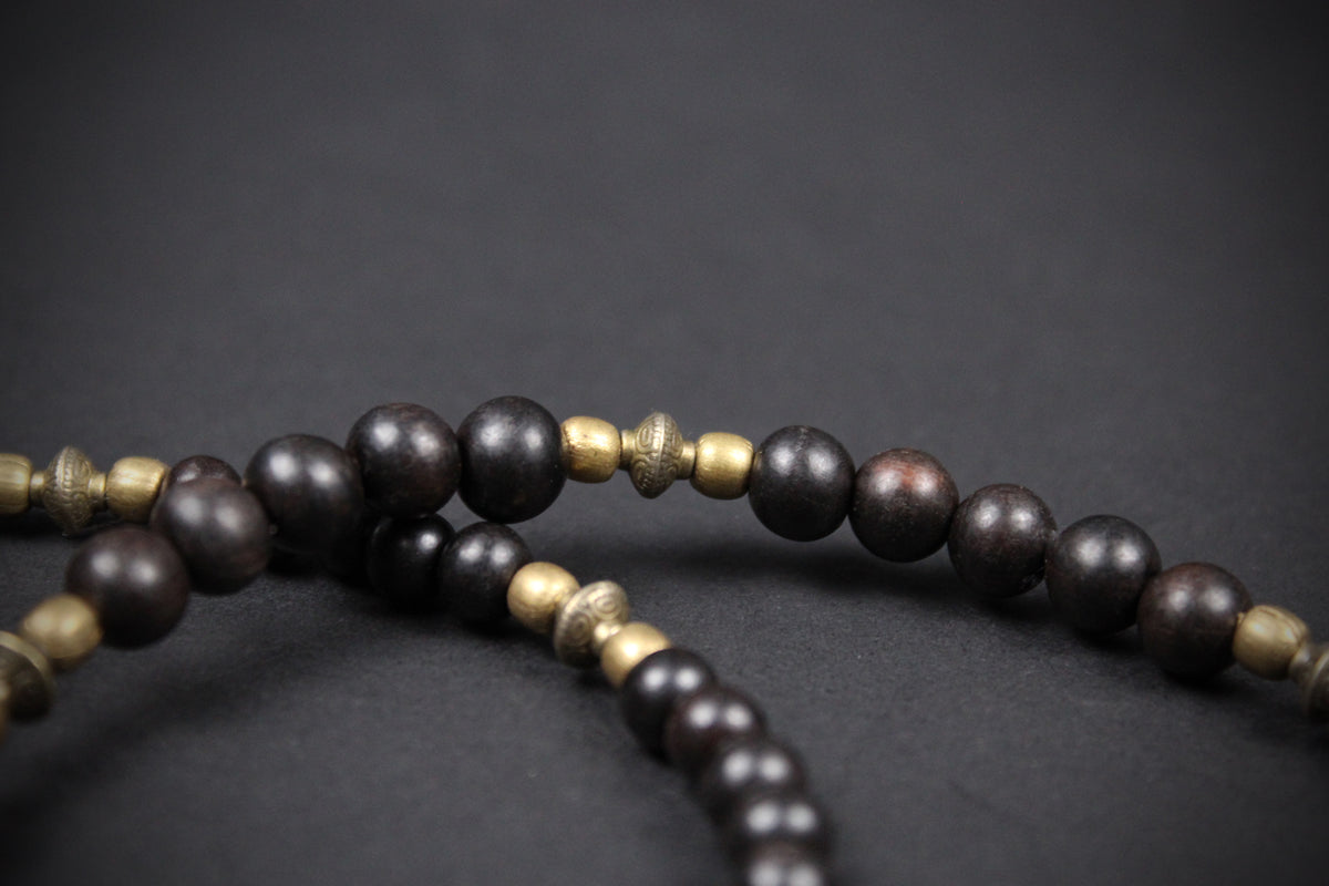 Dark brown wooden prayer beads with small metallic gold spacer beads, shown in a close-up shot against a dark gray background.