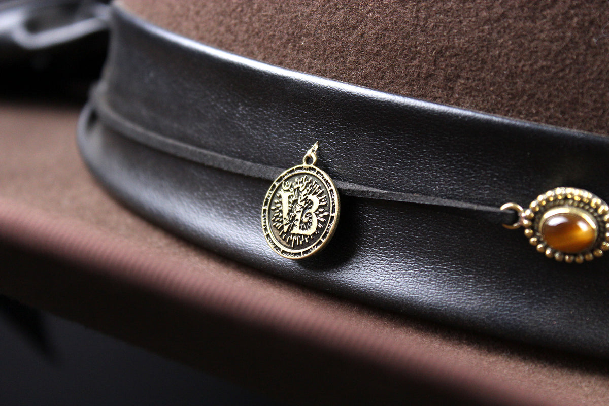 A close-up of a brown felt hat with a black leather band decorated with a round gold-toned medallion and a gold-toned concho-style ornament.