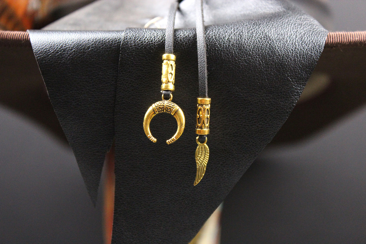 A pair of gold-toned pendant necklaces with engraved cylindrical beads and a crescent moon and wing charms, hanging from black cords against a black textured display stand.
