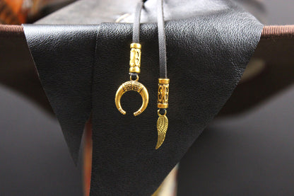 A pair of gold-toned pendant necklaces with engraved cylindrical beads and a crescent moon and wing charms, hanging from black cords against a black textured display stand.