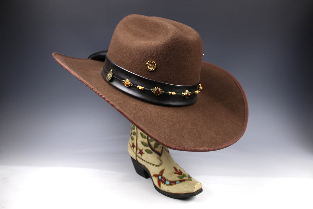A brown felt cowboy hat with a black leather band adorned with gold-toned studs and medallions, displayed on a decorative boot-shaped stand against a dark gray studio background.