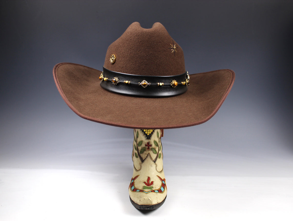 A brown felt cowboy hat with a black band decorated with gold-toned studs and a floral embroidered boot displayed underneath on a dark gray gradient background.