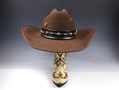 A brown felt cowboy hat with a black band decorated with gold-toned studs and a floral embroidered boot displayed underneath on a dark gray gradient background.