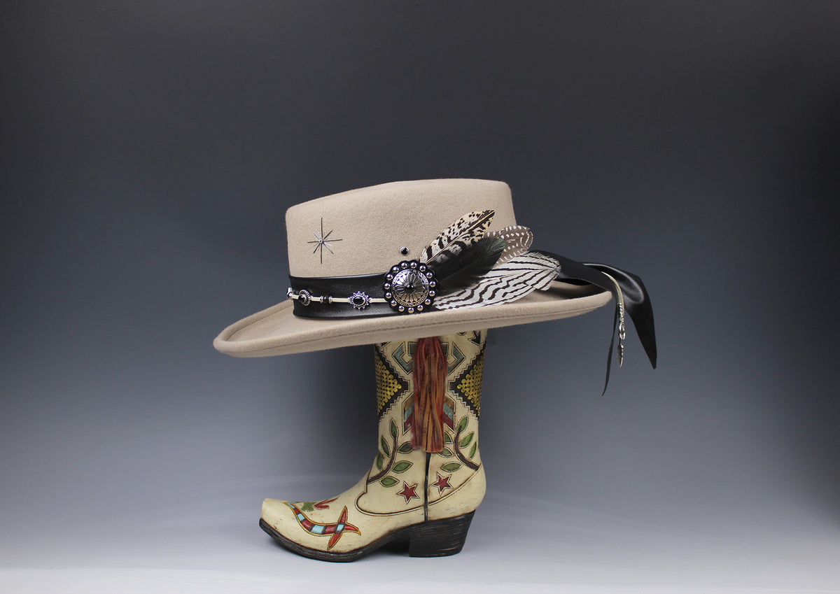 A beige wide-brim cowboy hat with a black band, feather, beadwork, and tassel, displayed on top of a decorated cream western boot against a dark gray gradient background.