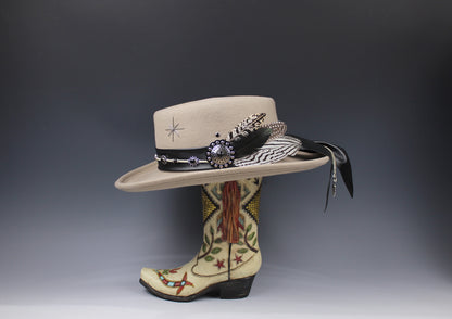 A beige wide-brim cowboy hat with a black band, feather, beadwork, and tassel, displayed on top of a decorated cream western boot against a dark gray gradient background.