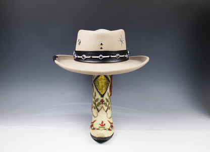 A beige cowboy hat with a black beaded band, displayed on top of a decorated cowboy boot against a dark gray gradient background.