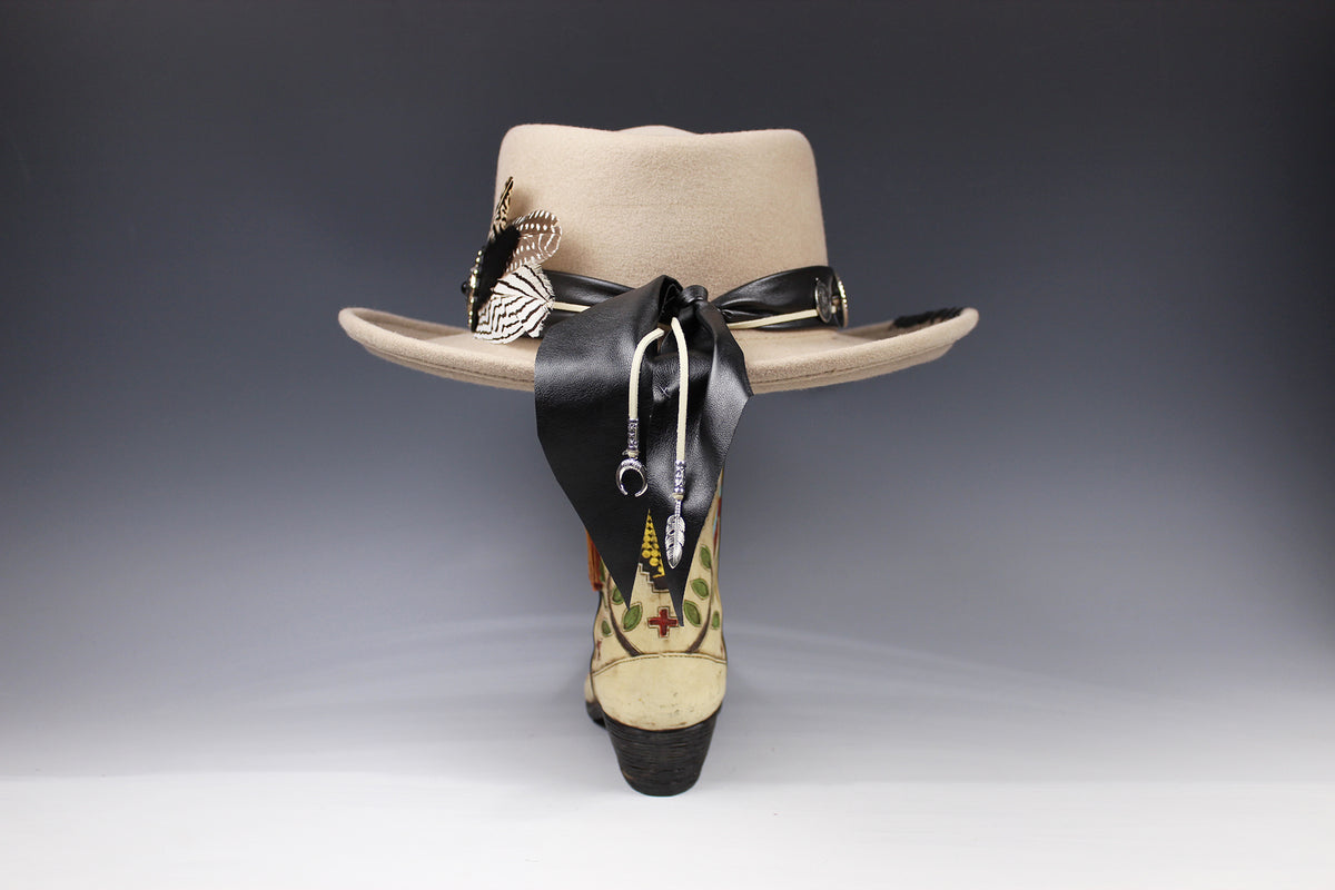 A beige wide-brim fedora hat displayed from the back, featuring a black ribbon tied in a bow, silver-toned decorative pins, and a highly embroidered heel in beige, black, and green.