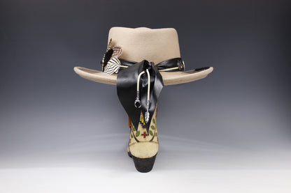 A beige wide-brim fedora hat displayed from the back, featuring a black ribbon tied in a bow, silver-toned decorative pins, and a highly embroidered heel in beige, black, and green.