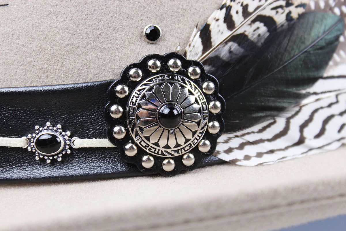 Close-up of a western-style hat band in black leather with a large silver concho-style ornament, small silver studs, a black bead, and a dark feather with a black-and-white patterned base on a beige hat.
