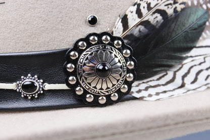 Close-up of a western-style hat band in black leather with a large silver concho-style ornament, small silver studs, a black bead, and a dark feather with a black-and-white patterned base on a beige hat.