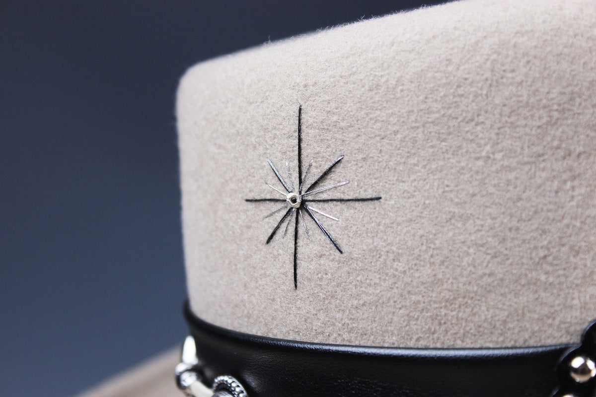 Close-up of a beige felt military-style cap with a black leather band and a silver starburst emblem on the front, set against a dark blue-gray background.