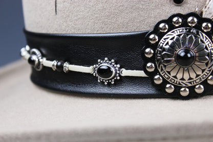 A close-up of a black leather belt with silver-toned metal hardware, including a large decorative medallion and smaller matching accents, displayed on a beige mannequin stand.