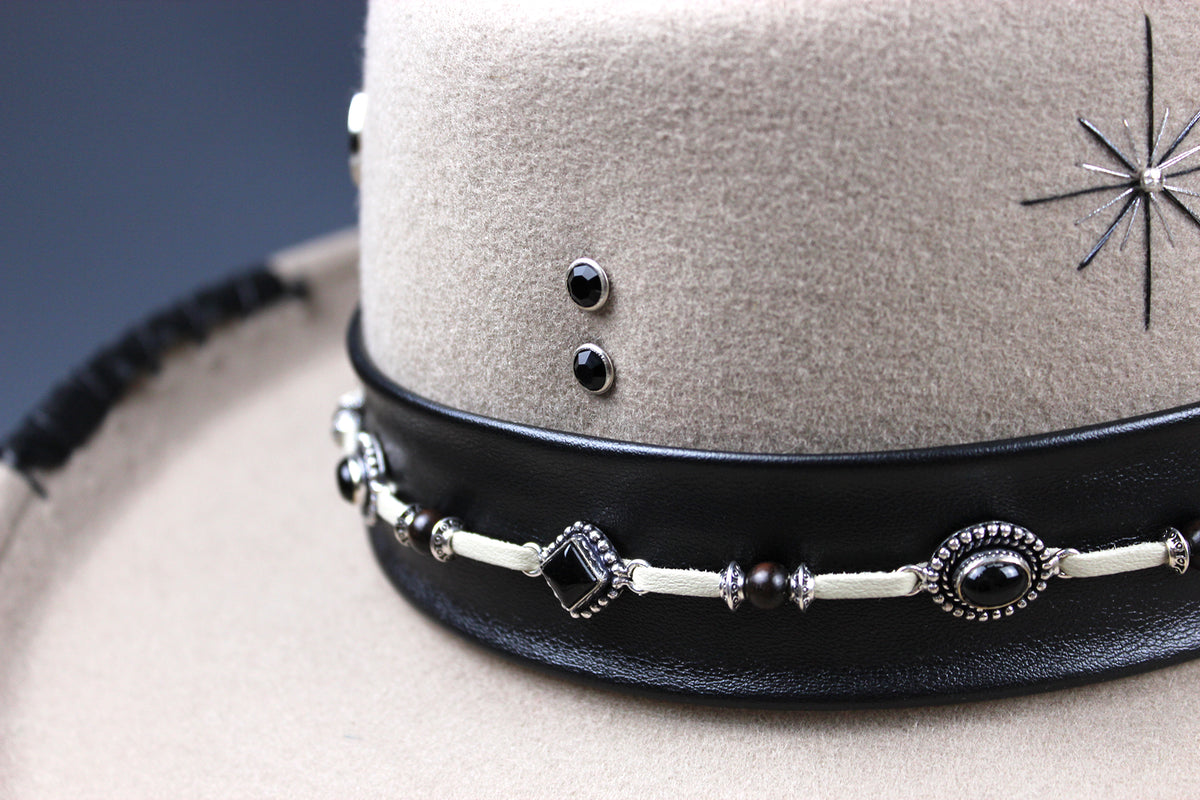 Close-up of a beige felt hat with a wide black leather band decorated with silver-toned beads, dark stones, and a starburst emblem.