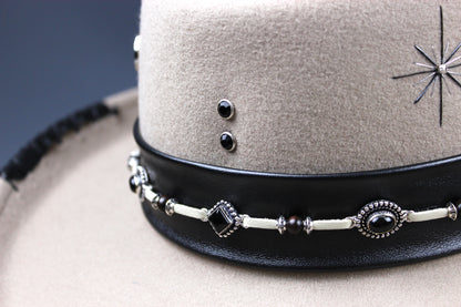 Close-up of a beige felt hat with a wide black leather band decorated with silver-toned beads, dark stones, and a starburst emblem.