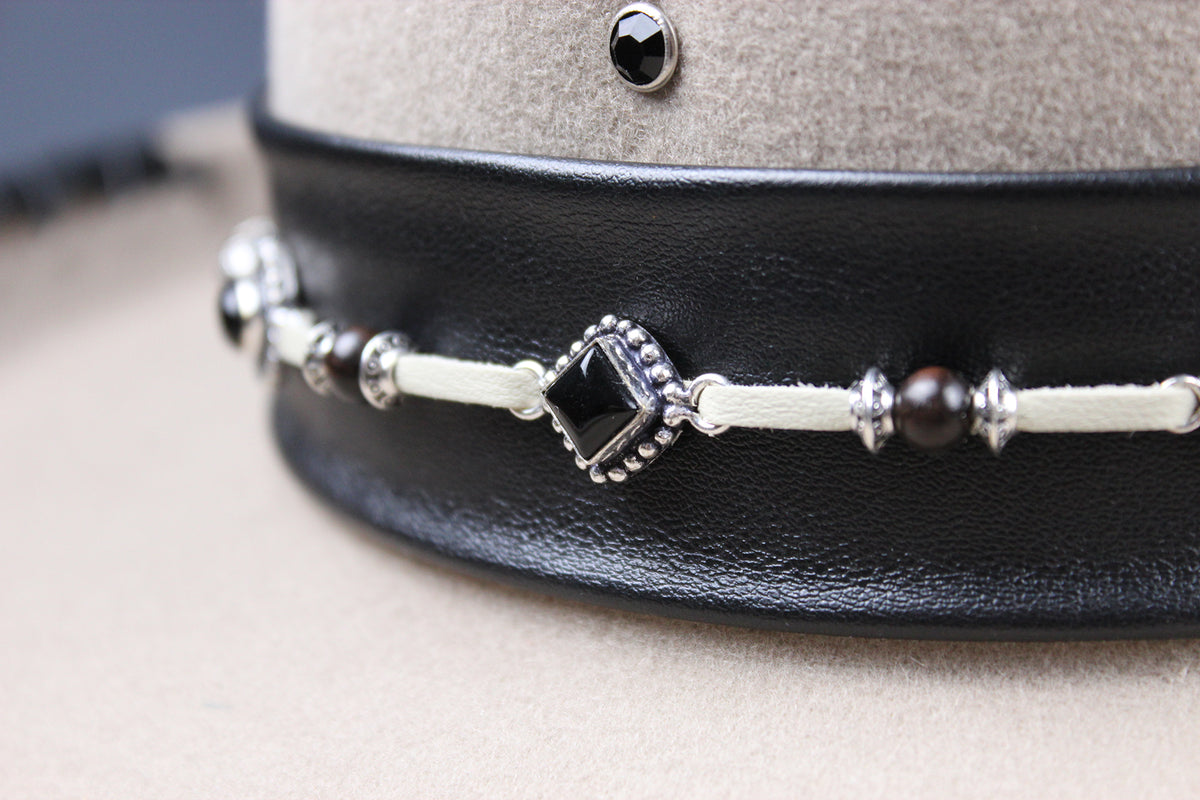 A close-up of a black leather choker collar featuring a decorative white cord bracelet with a central black diamond-shaped charm, silver-toned beads, and dark brown beads, displayed on a beige mannequin stand.