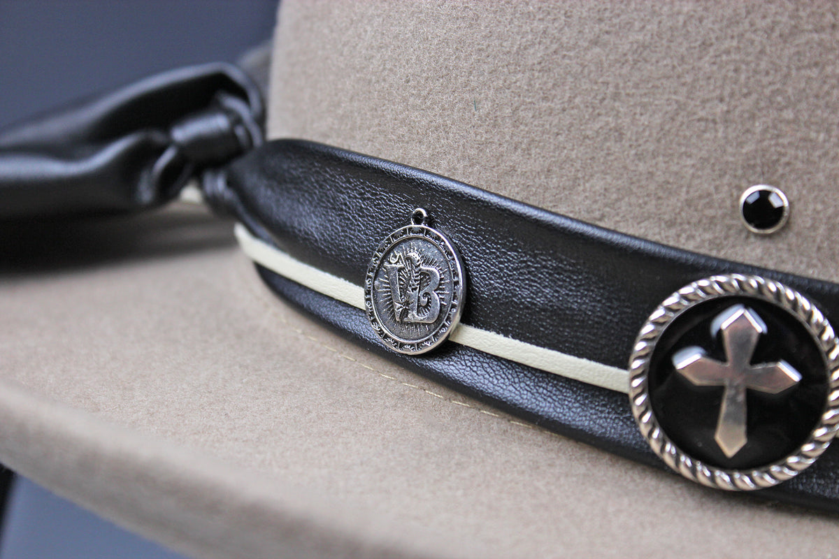 Close-up of a beige cowboy hat featuring a black leather band with a white stripe, a silver circular medallion, and a large silver cross concho.